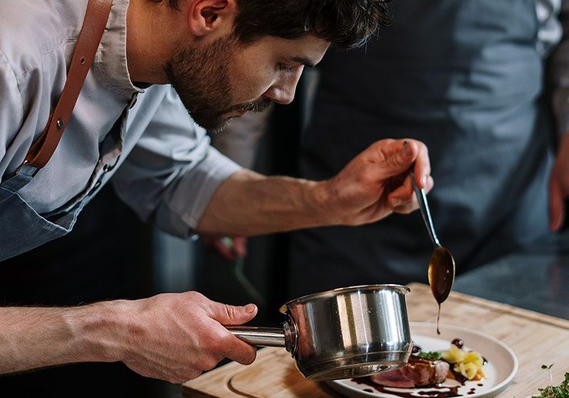 A chef preparing a dish and putting a liquid over the dish.